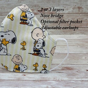 May include: A face mask featuring a repeating pattern of Snoopy, Charlie Brown, and Woodstock characters on a light yellow striped background. The mask has white ear loops and text that reads: 2 or 3 layers, Nose bridge, Optional filter pocket, Adjustable earloops.