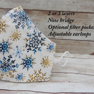 May include: A white face mask with a blue and gold snowflake pattern. The mask has a nose bridge, adjustable ear loops, and an optional filter pocket. 2 or 3 layers.