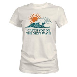 May include: White t-shirt featuring a graphic of a sun and a wave in shades of orange and teal. The text "CATCH YOU ON THE NEXT WAVE" is printed below the graphic. The shirt has short sleeves and a crew neck.