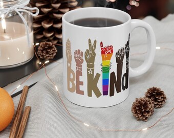 May include: White ceramic mug with a design of hands in different skin tones forming the sign language for "be kind". The hands are decorated with rainbow colors and stars.
