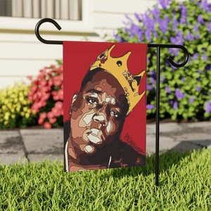 May include: A red garden flag with a cartoon illustration of a man wearing a gold crown. The man's face is detailed and the background is red. The text "King" is visible in the bottom right corner of the flag.