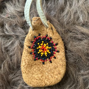 Puede incluir: Una pequeña bolsa de cuero marrón claro con un diseño de sol de cuentas en rojo, negro y amarillo. La bolsa tiene un cordón de cuero color crema para llevar alrededor del cuello. El artículo es hecho a mano.