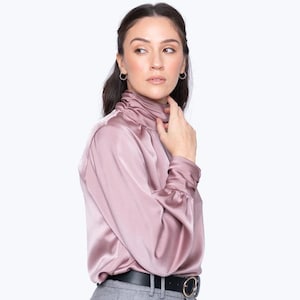 Könnte beinhalten: Eine mauvefarbene Satinbluse mit hohem, gerüschtem Ausschnitt und langen Ärmeln. Die Bluse wird mit einer grauen Hose und einem schwarzen Gürtel mit goldener Schnalle getragen. Das Model trägt kleine silberne Creolen.