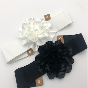 May include: Two elastic waist belts, one white and one black, each featuring a large, fabric flower. The belts have brown leather-like accents and metal snap closures. A stylish accessory for dresses and tops.
