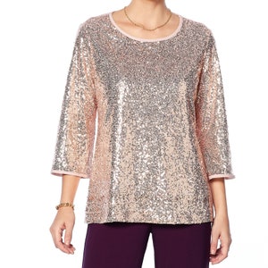 Può includere: Top di paillettes rosa chiaro con maniche a tre quarti. Il top ha uno scollo tondo e un bordo rosa sulle maniche e sullo scollo. Sono visibili anche una collana e un braccialetto color oro.