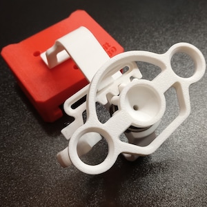 May include: White 3D printed phone holder with a red base. The holder has a circular design with a hole in the center and two side loops. The base has a small hole on the top and two holes on the side.