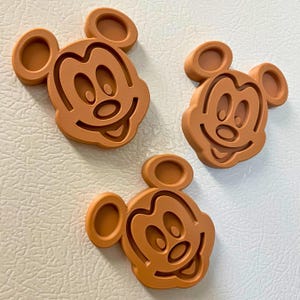 May include: Three brown Mickey Mouse-shaped magnets. Each magnet features the iconic character's face with large ears, a smiling mouth, and oval eyes. The magnets are made of a solid material and are placed on a white surface.