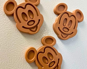 May include: Three brown Mickey Mouse-shaped magnets. Each magnet features the iconic character's face with large ears, a smiling mouth, and oval eyes. The magnets are made of a solid material and are placed on a white surface.