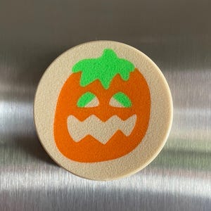 May include: A round, beige-colored, ceramic coaster with an orange pumpkin face with a green stem and eyes. The pumpkin has a toothy grin.