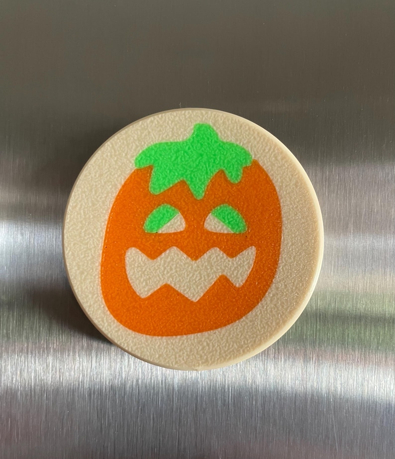 May include: A round, beige-colored, ceramic coaster with an orange pumpkin face with a green stem and eyes. The pumpkin has a toothy grin.
