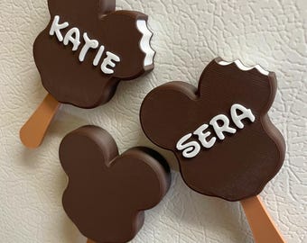 May include: Three brown Mickey Mouse-shaped ice cream bar magnets. Two have white text that reads "KATIE" and "SERA". One has a bite taken out of it. Each magnet has a light brown popsicle stick.