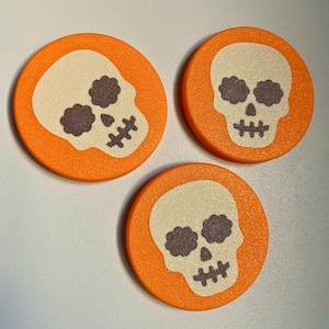 May include: Three circular orange discs, each featuring a cream-colored skull graphic with dark brown, flower-shaped eye sockets and a stitched mouth design. The surface of each disc has a subtle, uniform texture throughout the entire design.