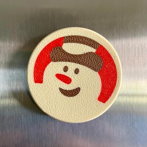 May include: A round, off-white Christmas-themed magnet featuring a smiling snowman face. The snowman has a brown hat, a red scarf, and a red nose. The magnet is likely made of plastic and has a textured surface.