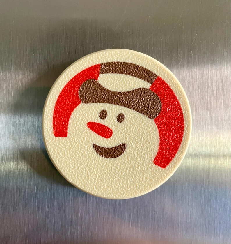 May include: A round, off-white Christmas-themed magnet featuring a smiling snowman face. The snowman has a brown hat, a red scarf, and a red nose. The magnet is likely made of plastic and has a textured surface.