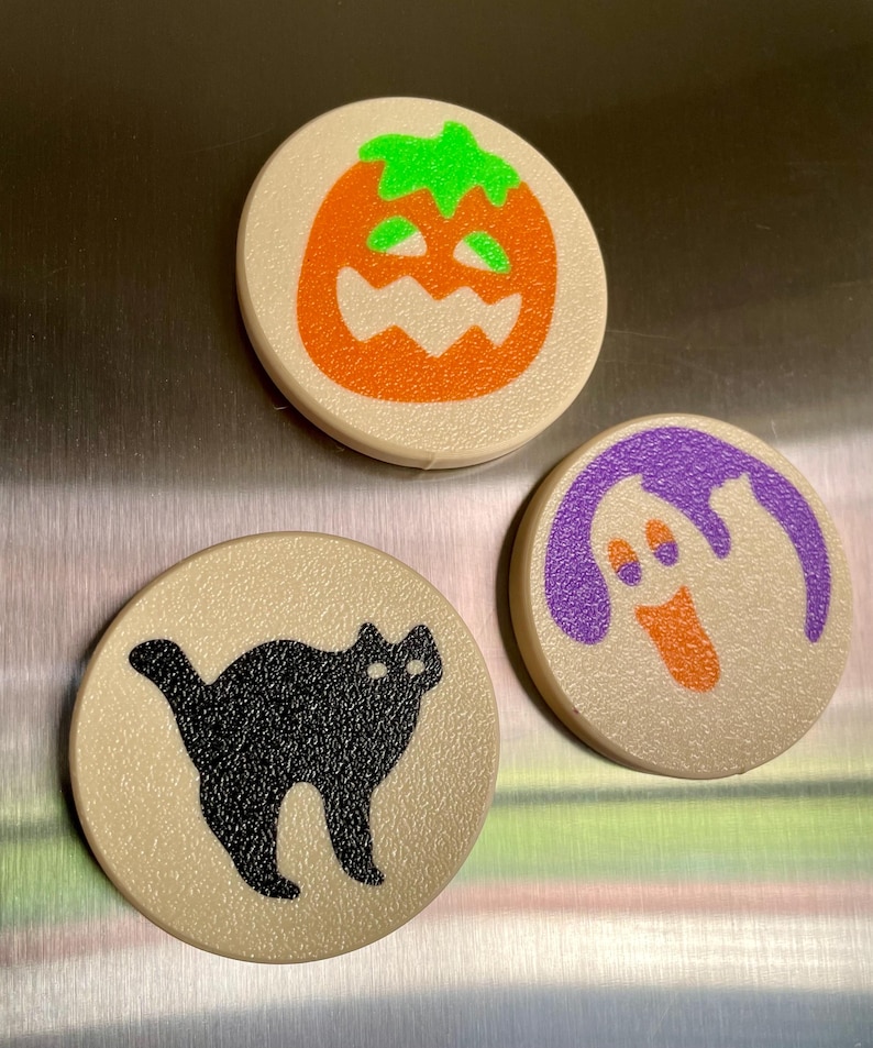 May include: Three round Halloween themed magnets. The first magnet is beige with an orange pumpkin with a green stem and a jack-o-lantern face. The second magnet is beige with a black cat silhouette. The third magnet is beige with a purple ghost silhouette with an orange face.