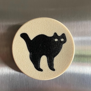 May include: A round, cream-colored magnet with a black silhouette of a cat with white eyes.