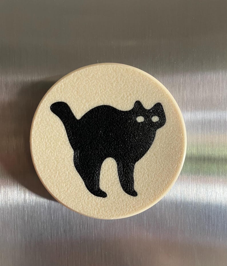 May include: A round, cream-colored magnet with a black silhouette of a cat with white eyes.