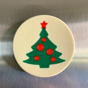 May include: A round, off-white Christmas tree magnet. The green tree has red ornaments and a red star on top. The magnet is a festive holiday decoration.