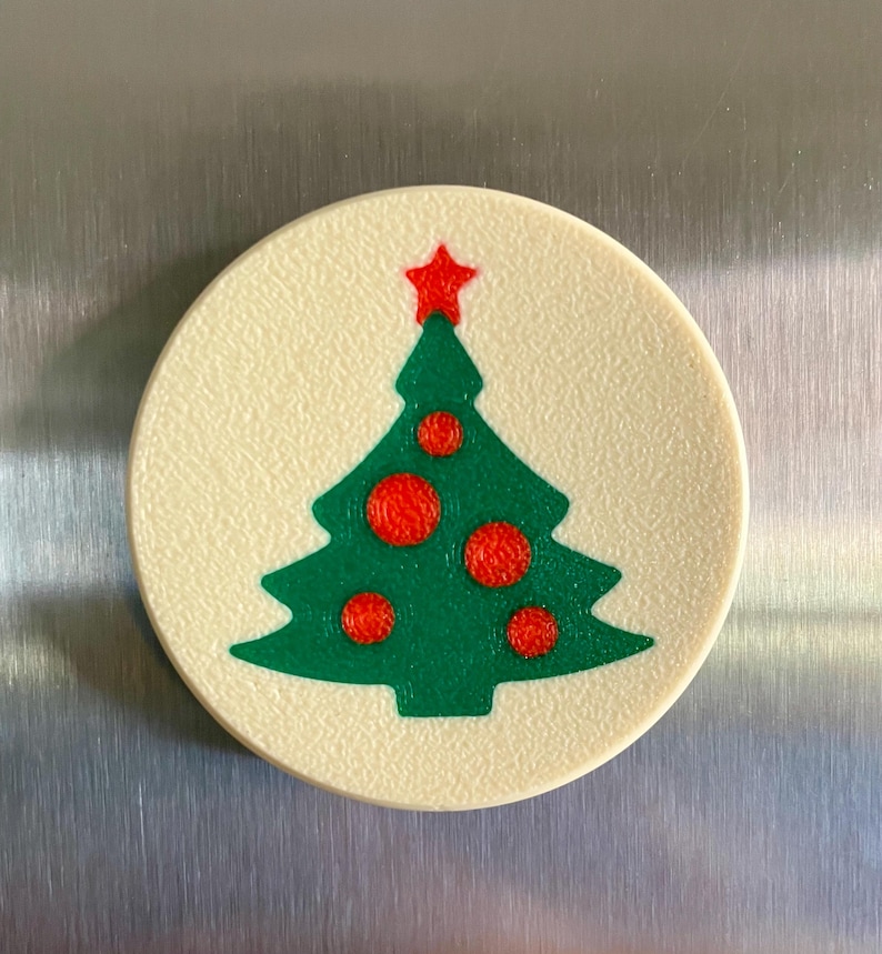May include: A round, off-white Christmas tree magnet. The green tree has red ornaments and a red star on top. The magnet is a festive holiday decoration.