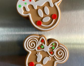 May include: Two gingerbread cookie magnets shaped like Mickey Mouse and Minnie Mouse. The magnets are decorated with white icing, red and green sprinkles, and a white bow on Minnie Mouse.