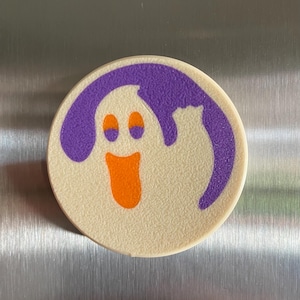 May include: A white round magnet with a purple outline and a cartoon ghost with orange eyes and mouth. The ghost is smiling.