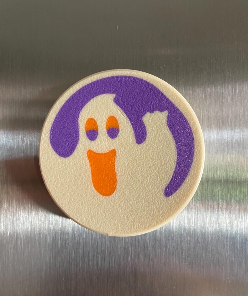 May include: A white round magnet with a purple outline and a cartoon ghost with orange eyes and mouth. The ghost is smiling.