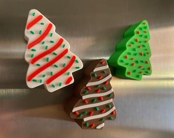 May include: Three Christmas tree-shaped magnets. One is white with red stripes and green sprinkles, another is green with red sprinkles, and the third is brown with white stripes and green sprinkles. The magnets are on a silver surface.