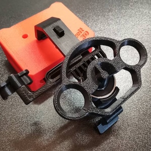 May include: A 3D printed black camera mount with a red 3D printed case attached to it. The mount has a circular design with three holes and a small knob. The case has a small hole and a textured surface.