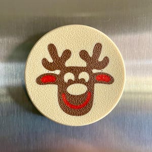 May include: A round, off-white magnet with a textured surface. The magnet features a cartoon reindeer design with brown antlers, a brown face, red nose and ears, and a red smiling mouth. The magnet is likely for a refrigerator.