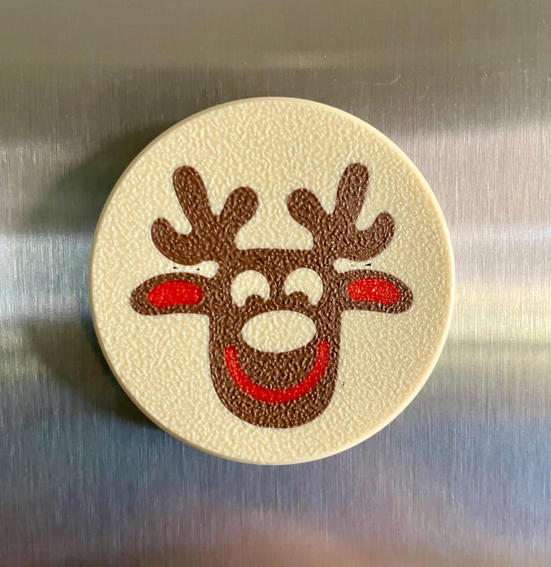 May include: A round, off-white magnet with a textured surface. The magnet features a cartoon reindeer design with brown antlers, a brown face, red nose and ears, and a red smiling mouth. The magnet is likely for a refrigerator.