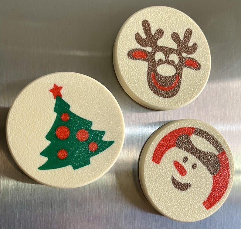 May include: Three round Christmas-themed magnets. One features a green Christmas tree with red ornaments and a red star. Another depicts a reindeer with brown antlers and a red nose. The last shows a snowman with a red hat.