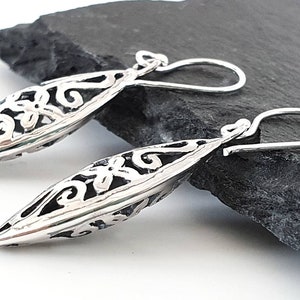 May include: A pair of silver dangle earrings with a delicate, intricate design. The earrings are shaped like a long, narrow oval with a floral pattern.