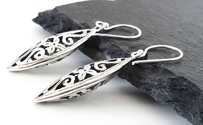 May include: A pair of silver dangle earrings with a delicate, intricate design. The earrings are shaped like a long, narrow oval with a floral pattern.