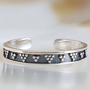 Pode incluir: Anel aberto prateado com uma faixa cinza escuro. A faixa é decorada com pequenos pontos prateados em relevo, dispostos em grupos de três. O anel tem um design simples e elegante.