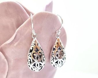 May include: A pair of silver filigree teardrop earrings with a delicate, intricate design. The earrings are hanging from a pink background.