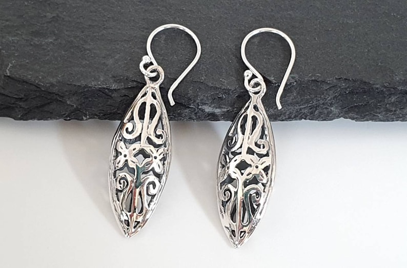 May include: A pair of silver dangle earrings with a delicate, intricate design. The earrings are oval-shaped and feature a floral pattern.
