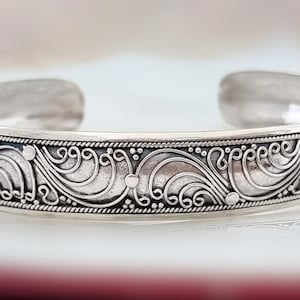 Könnte beinhalten: Ein silbernes Armreif-Armband mit offenem Design. Das Armband weist ein kompliziertes Muster aus wirbelnden Linien und Punkten auf, das einen detaillierten, ornamentalen Look erzeugt. Das Metall hat eine polierte, reflektierende Oberfläche. Das Armband wird auf einer weißen Oberfläche präsentiert.