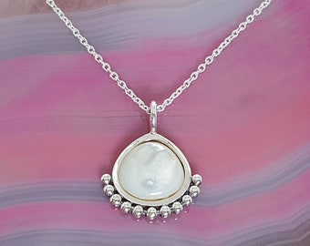 Op de afbeelding: Een zilveren ketting met een traanvormige hanger met een witte parelmoer inleg omringd door een rij zilveren kralen.