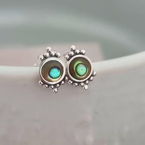 Può includere: Un paio di orecchini a lobo in argento. Ogni orecchino presenta un centro rotondo in conchiglia di abalone iridescente, circondato da una lunetta in argento e dettagli decorativi in argento. La conchiglia di abalone mostra sfumature di verde e blu.