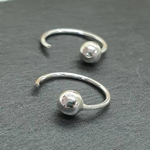 Op de afbeelding: Twee zilverkleurige, open oorringen. Elke oorring heeft een kleine, ronde bal aan één uiteinde. De oorbellen worden gepresenteerd op een donkergrijze ondergrond, wat hun eenvoudige, moderne ontwerp benadrukt. Sieraden.