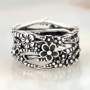 Könnte beinhalten: Ein silberner Ring mit einem floralen Design. Der Ring besteht aus mehreren Bändern, die miteinander verwoben sind und mit kleinen Blumen verziert sind.