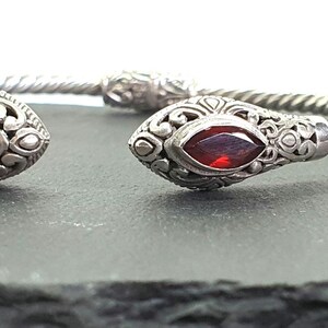 Pode incluir: Pulseira de punho prateada com detalhes de cabeça de cobra. Cada cabeça de cobra apresenta uma pedra preciosa vermelha escura com corte marquesa. A pulseira tem um design de corda torcida e é exibida em uma superfície escura.