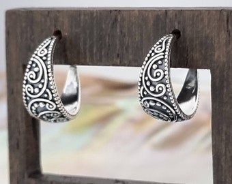 May include: A pair of silver hoop earrings with a detailed, swirling, and dotted design. The earrings are displayed on a wooden stand.