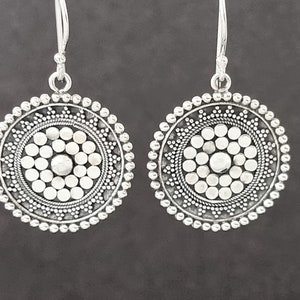 Könnte beinhalten: Ein Paar silberne Ohrringe mit einem runden Design. Die Ohrringe haben einen zentralen Kreis, der von kleineren Kreisen umgeben ist und ein Mandala-artiges Muster erzeugt.