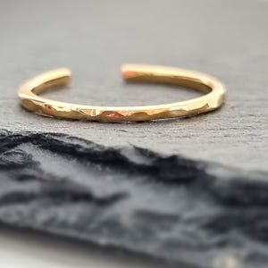 Könnte beinhalten: Ein goldfarbener offener Ring mit strukturierter Oberfläche. Der Ring liegt auf einem dunkelgrauen, strukturierten Hintergrund. Das Design des Rings weist ein gehämmertes oder facettiertes Aussehen auf und sorgt für visuelles Interesse.