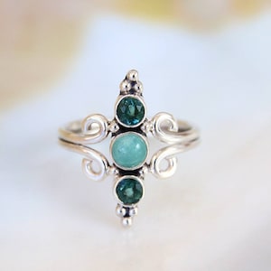 Puede incluir: Un anillo de plata con un diseño delicado que presenta tres piedras preciosas. La piedra central es de color verde claro, mientras que las dos piedras más pequeñas a cada lado son de color verde oscuro.