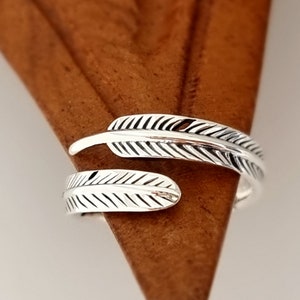 Pode incluir: Um anel ajustável prateado com um design de pena. O anel apresenta duas formas de pena detalhadas, com linhas gravadas que criam a textura da pena. O anel é mostrado em uma superfície marrom.