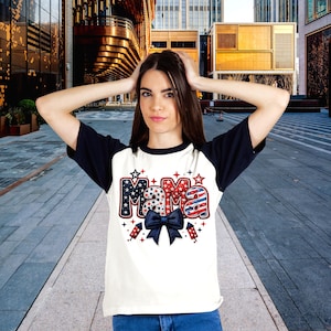 Puede incluir: Camiseta de béisbol blanca con mangas azul marino. La palabra "Mama" está impresa en rojo, blanco y azul, con estrellas, fuegos artificiales y un lazo azul marino.