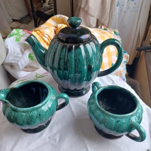 May include: A green glazed ceramic teapot with a matching creamer and sugar bowl. The teapot has a rounded body with a fluted design and a black handle. The creamer and sugar bowl have a similar fluted design.