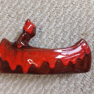 May include: A vintage ceramic ashtray shaped like a canoe. The ashtray is glazed in a glossy red and brown colour scheme. The canoe has a small figure on the back. The ashtray is sitting on a beige carpet.